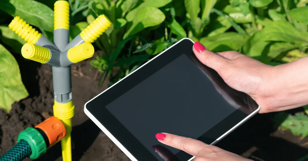 A hand reaching out to tap a tablet screen controlling an irrigation system. They hold the tablet above a field with a hose.