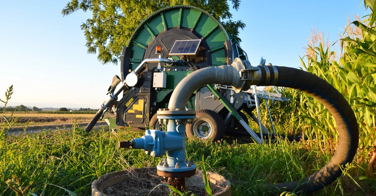 A water pump, up close, in a farm field, with a hose connected to a large, green hose reel in the background.