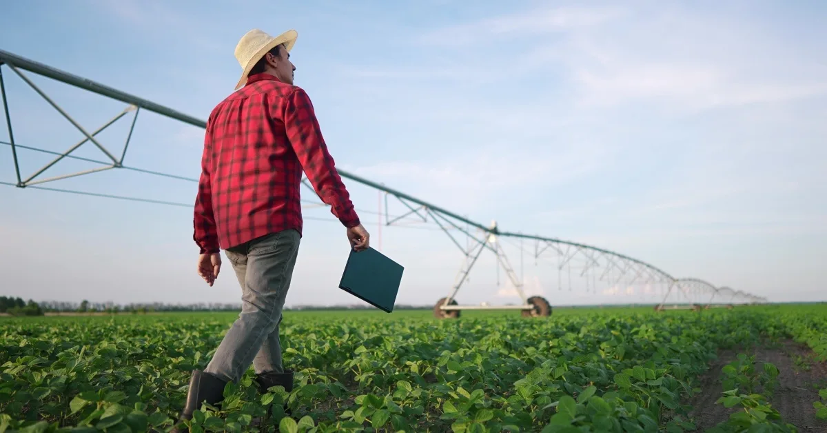 A male farmer in a red shirt and sun hat walking through a farm field with a tablet in-hand, looking up at the sky.