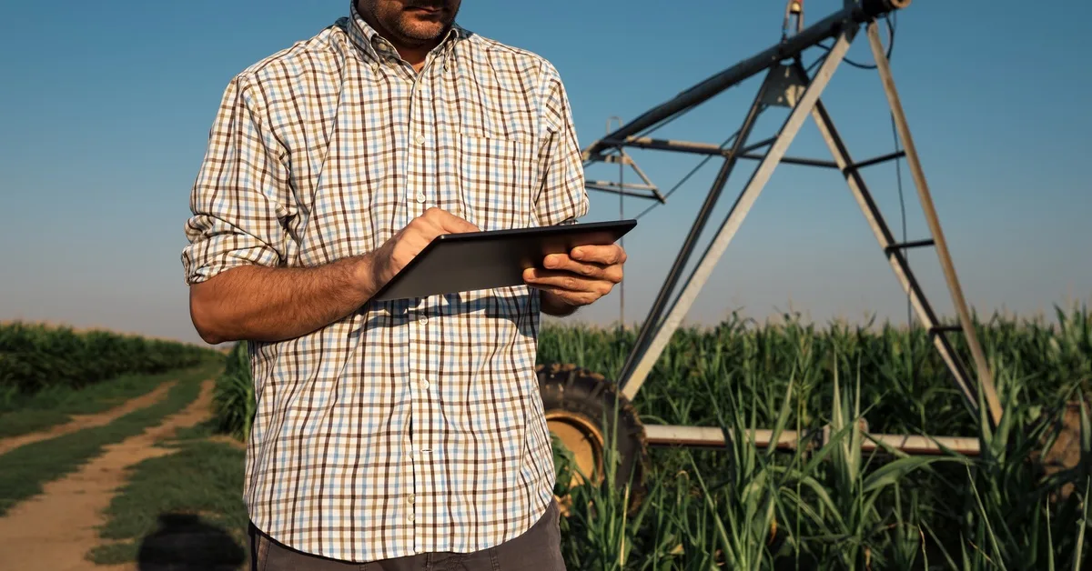 A close-up of a man's upper body holding a tablet with an irrigation system and corn field in the background.