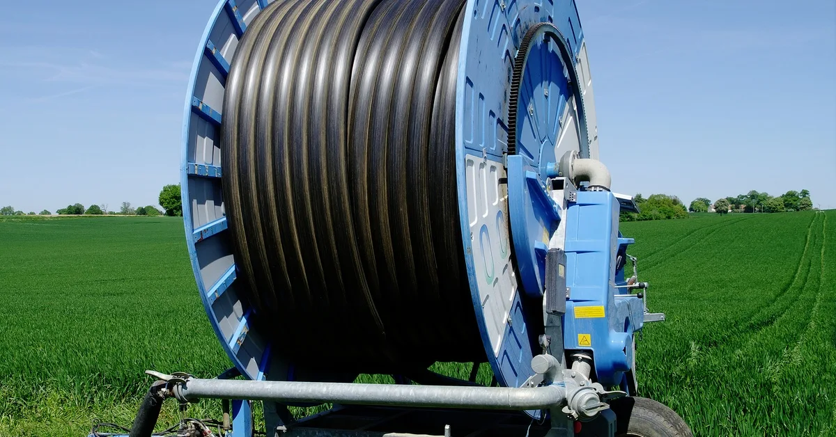 A large, blue hose reel with black hose running through it sits on the edge of a vast, green farm field. A large, blue hose reel with black hose running through it sits on the edge of a vast, green farm field.