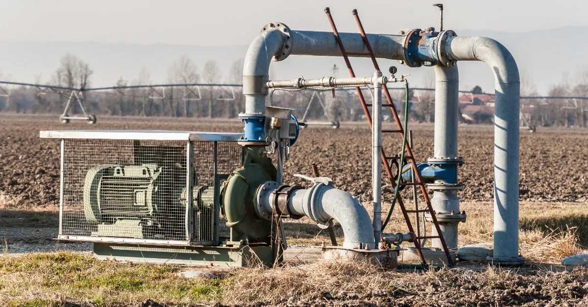 A large irrigation pump and metal pipes standing beside a plowed field with a ladder and sprinklers in the distance.